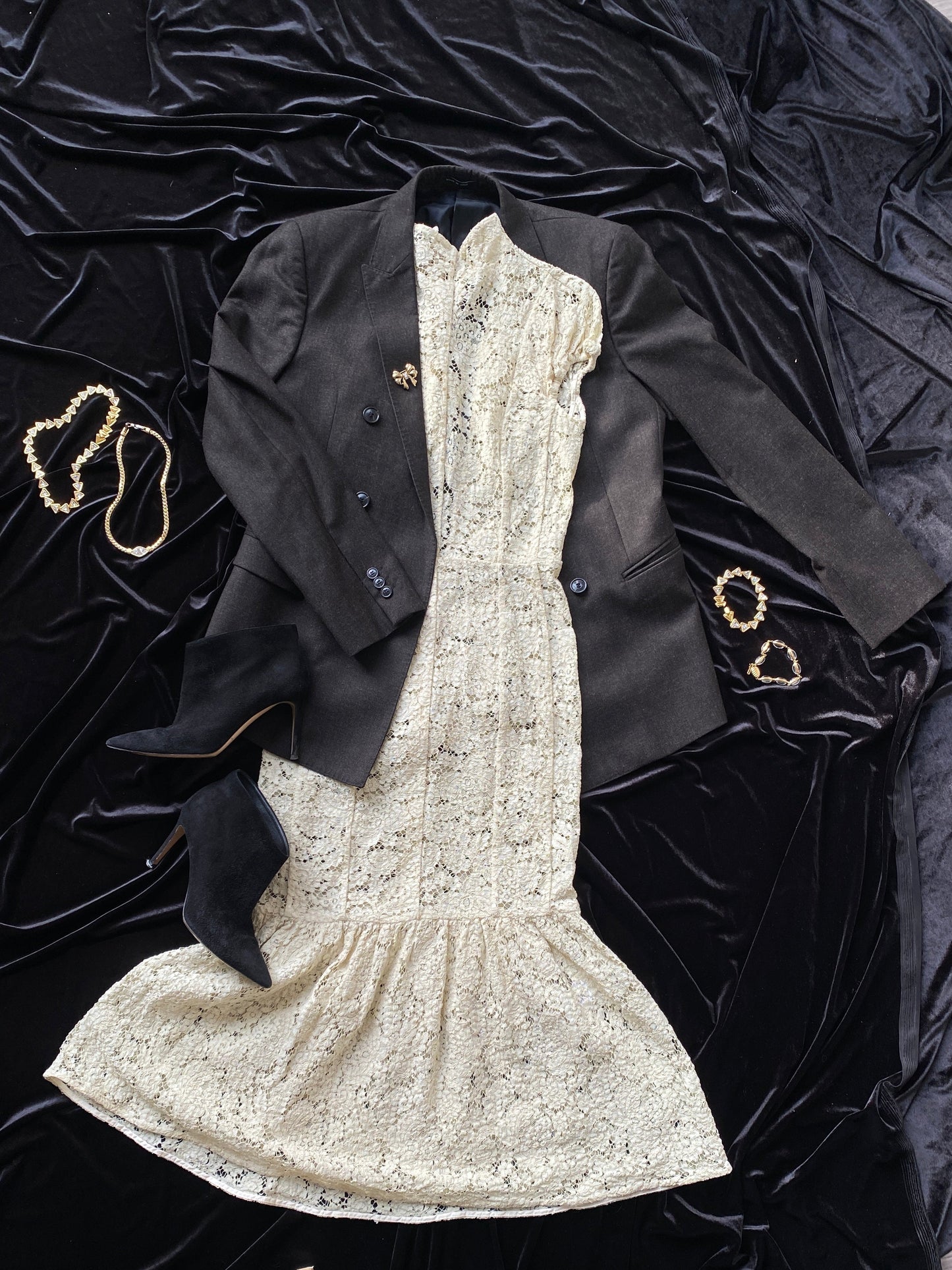A gray Giorgio Armani Double-Breasted Jacket lying on a black background. The jacket is partially placed over a white lace Victoria Beckham dress. Black boots sit beside, to the bottom left. Two necklaces are placed on the left side and two bracelets are placed on the right side.