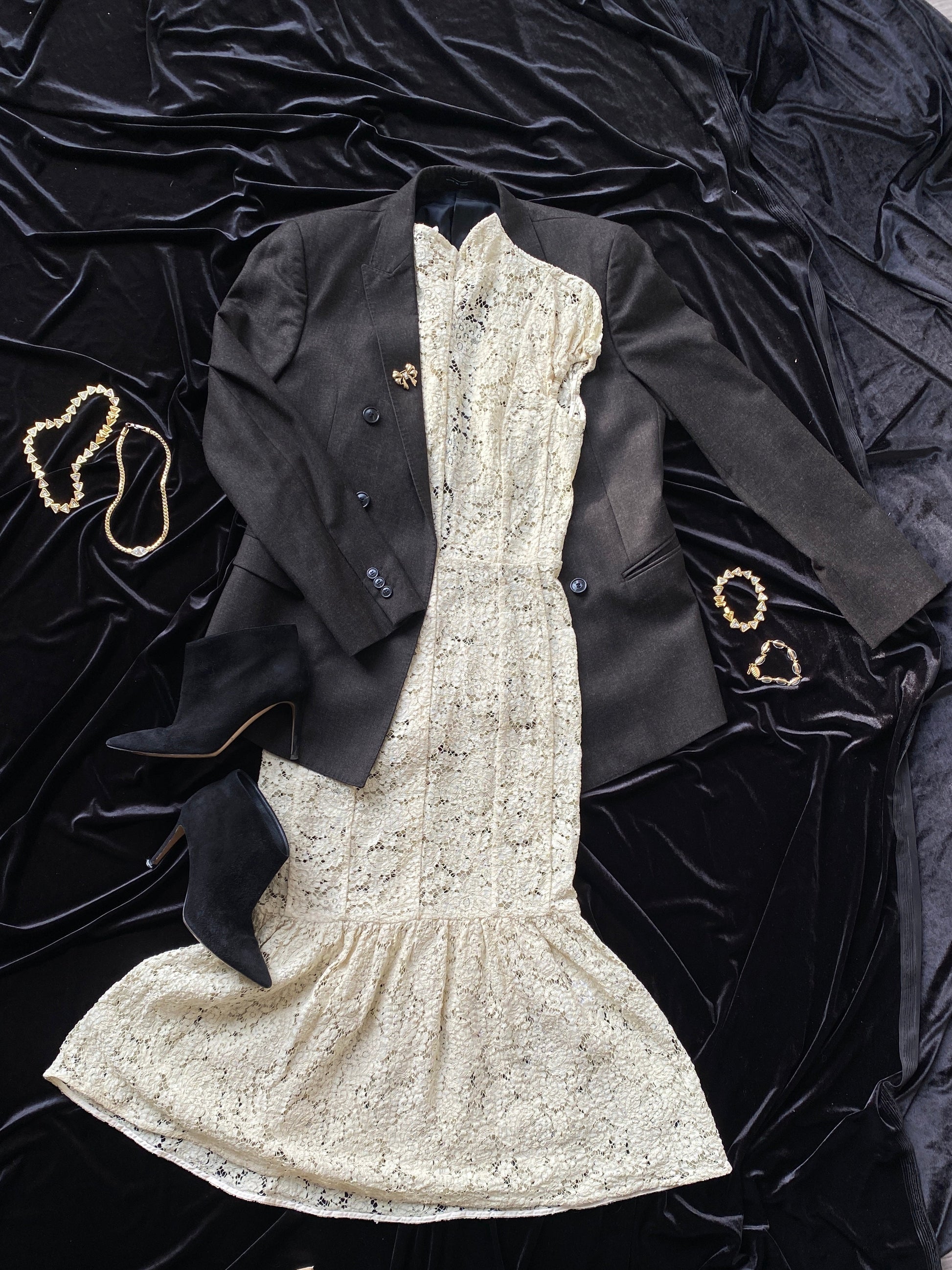 A gray Giorgio Armani Double-Breasted Jacket lying on a black background. The jacket is partially placed over a white lace Victoria Beckham dress. Black boots sit beside, to the bottom left. Two necklaces are placed on the left side and two bracelets are placed on the right side.