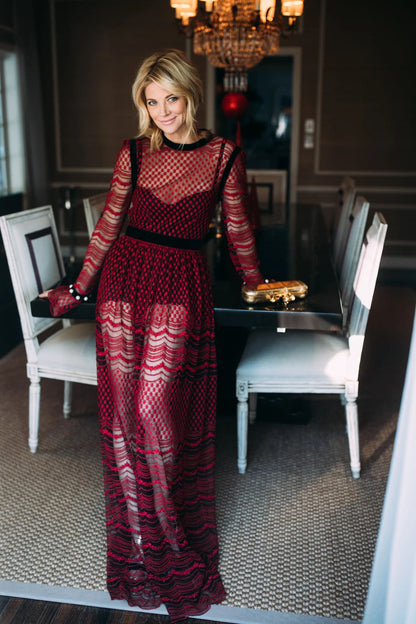 Woman modelling a Philosophy di Lorenzo Seraphini burgundy long sleeve gown dress standing in an elegant room, leaning on a table with a chandelier in the background.